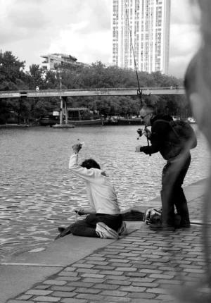 Pascale · 03/05/2026 · 1047 x 1500 Cette photographie en noir et blanc capture deux hommes près d’un plan d’eau. Le sujet principal semble être le personnage assis, en position de repos ou de méditation, tandis que l’autre, debout, semble l’observer ou interagir avec lui.La composition est dynamique, avec un cadrage serré sur les deux personnages. L’arrière-plan montre un pont et des bâtiments, ajoutant une touche urbaine. La lumière est naturelle, douce, créant des contrastes modérés. Les tons dominants sont les gris et noirs, typiques du noir et blanc, renforçant une ambiance contemplative et mélancolique. Le style est documentaire et spontané, évoquant une scène de vie quotidienne.