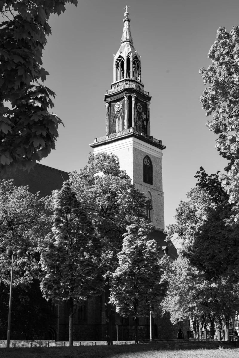 Claude · 3840 x 5760 Cette photographie met en scène un clocher d’église comme sujet principal, encadré par des arbres feuillus. La composition est symétrique, avec le clocher centré et les branches des arbres formant une sorte de cadre naturel.Le cadrage est serré sur la base du clocher, avec une perspective ascendante. La lumière est naturelle, probablement en journée, créant des contrastes nets entre ombres et lumières.En noir et blanc, les couleurs dominantes sont les nuances de gris, renforçant les textures et les jeux d’ombres. Le style est classique et épuré, évoquant une ambiance paisible et intemporelle.