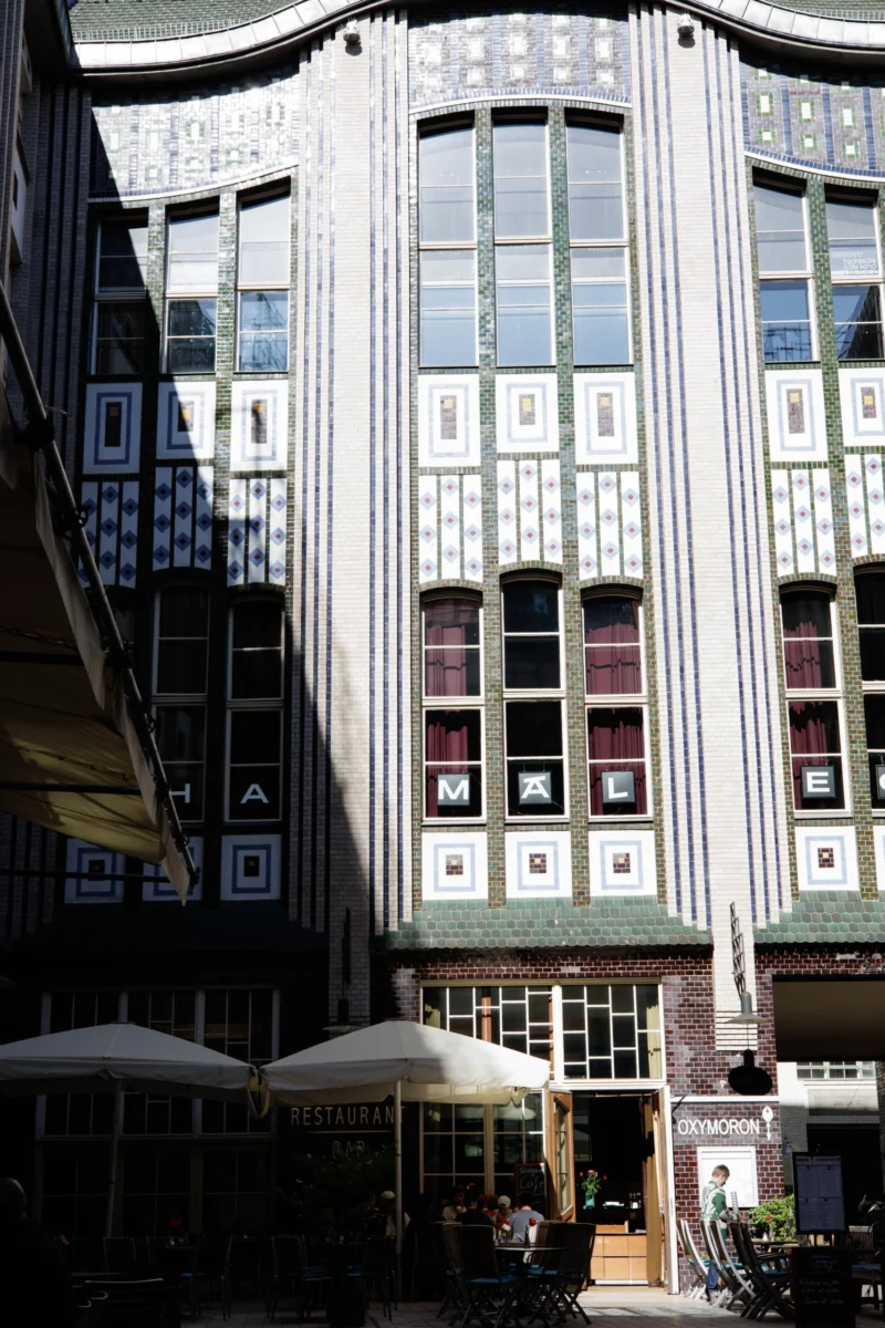 Claude · 3854 x 5781 Cette photographie montre un immeuble de style architectural Art déco ou moderne, avec des façades géométriques et des fenêtres alignées symétriquement. Le sujet principal est ce bâtiment, mis en valeur par un cadrage serré.La lumière naturelle éclaire la scène, créant des contrastes entre ombres et zones lumineuses. Les couleurs dominantes sont des tons neutres (beige, gris) avec des touches de blanc et de bleu.L’ambiance est à la fois urbaine et élégante, renforcée par les parasols et les tables à l’entrée, suggérant un espace public ou commercial.