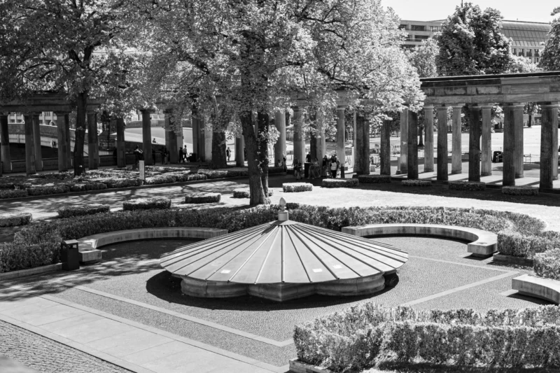 Claude · 5760 x 3840 Cette photographie en noir et blanc met en scène un **jardin urbain** avec pour **sujet principal** une fontaine circulaire centrale entourée de massifs fleuris bien entretenus.La **composition** est symétrique, avec des allées bordées d’arbres alignés et des bâtiments en arrière-plan. Le **cadrage** est large, offrant une vue panoramique.La **lumière** semble naturelle, douce, créant des contrastes nets entre ombres et clartés. Les **couleurs dominantes** sont les nuances de gris, accentuant les textures et les formes architecturales.Le **style** est classique et épuré, évoquant une **ambiance calme et ordonnée**, typique des espaces publics historiques.