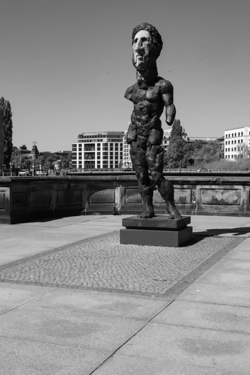 Claude · 3819 x 5729 Cette photographie montre une statue en bronze comme sujet principal, représentant un homme musclé et nu, debout sur un socle. La composition est épurée, avec un cadrage serré sur la statue, mettant en valeur ses détails. L’arrière-plan, flou, révèle un environnement urbain avec des bâtiments et un pont.La lumière est naturelle, contrastée, créant des ombres marquées. Les couleurs dominantes sont les tons gris et noirs, typiques d’une photo en noir et blanc, renforçant le côté dramatique et intemporel. Le style est réaliste et artistique, évoquant une ambiance à la fois moderne et introspective.