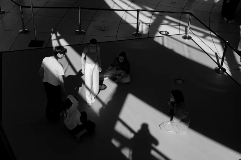 Claude · 6000 x 4000 Cette photographie en noir et blanc capture des silhouettes dans un espace intérieur moderne. Le sujet principal semble être les interactions entre les personnes, dont les ombres sont projetées sur le sol.La composition utilise un cadrage aérien, offrant une vue plongeante. La lumière naturelle, filtrée par des sources extérieures, crée des contrastes marqués et des jeux d'ombres dynamiques. Les couleurs dominantes sont les tons de gris, renforçant l'atmosphère dramatique et mystérieuse.Le style est épuré et contemporain, évoquant une ambiance à la fois calme et intrigante, presque cinématographique.
