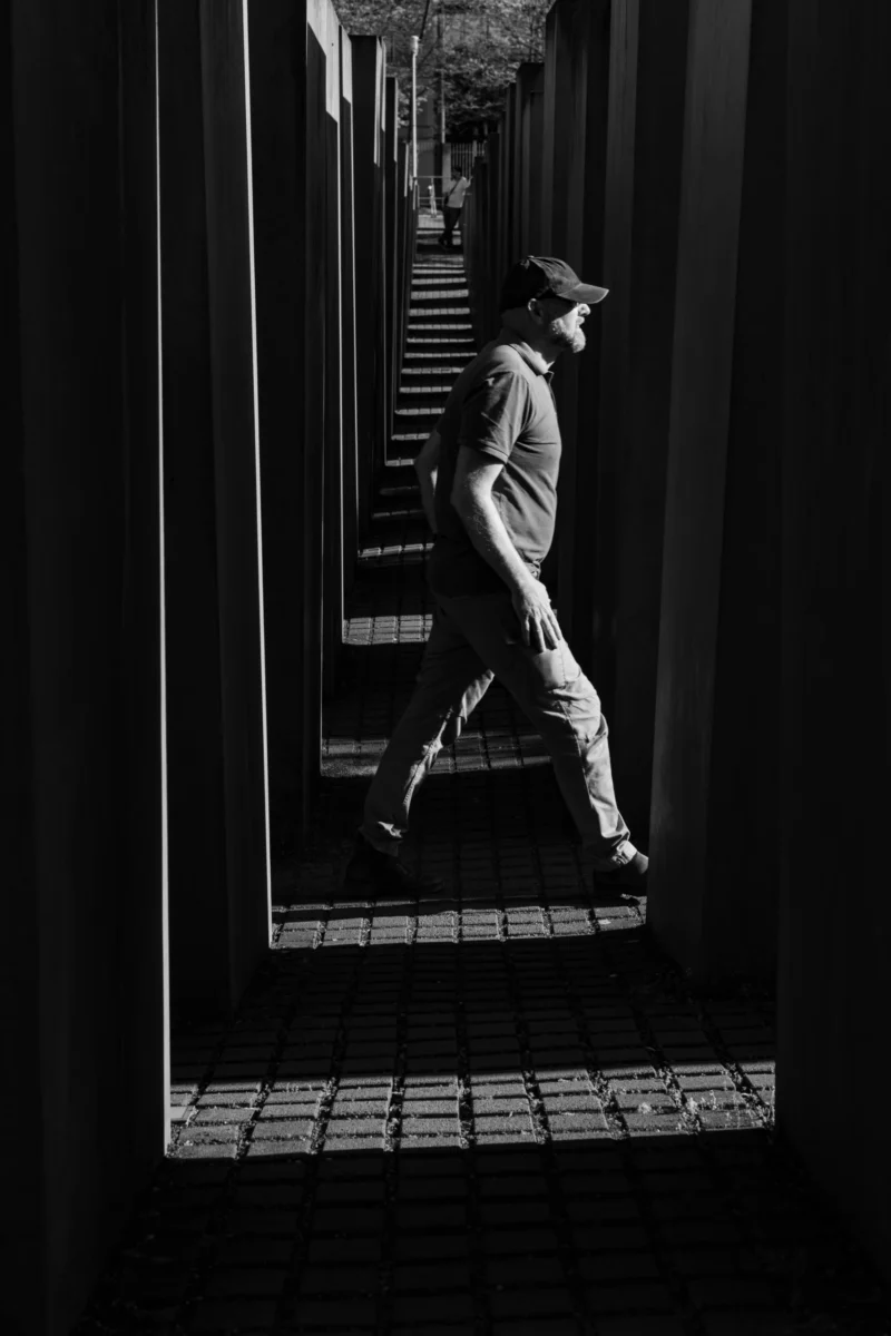 Claude · 3829 x 5743 Cette photographie en noir et blanc met en scène un homme comme sujet principal, marchant d’un pas vif dans un couloir étroit. La composition joue sur les lignes verticales et horizontales, créant un effet de profondeur et de perspective. Le cadrage serré isole le sujet, accentuant son mouvement.La lumière, contrastée, souligne les ombres et les formes, renforçant une ambiance dramatique. Les couleurs dominantes sont les nuances de gris, typiques du noir et blanc, avec des contrastes marqués entre clairs et obscurs. Le style est sobre et cinématographique, évoquant une atmosphère à la fois moderne et intemporelle, presque mélancolique.