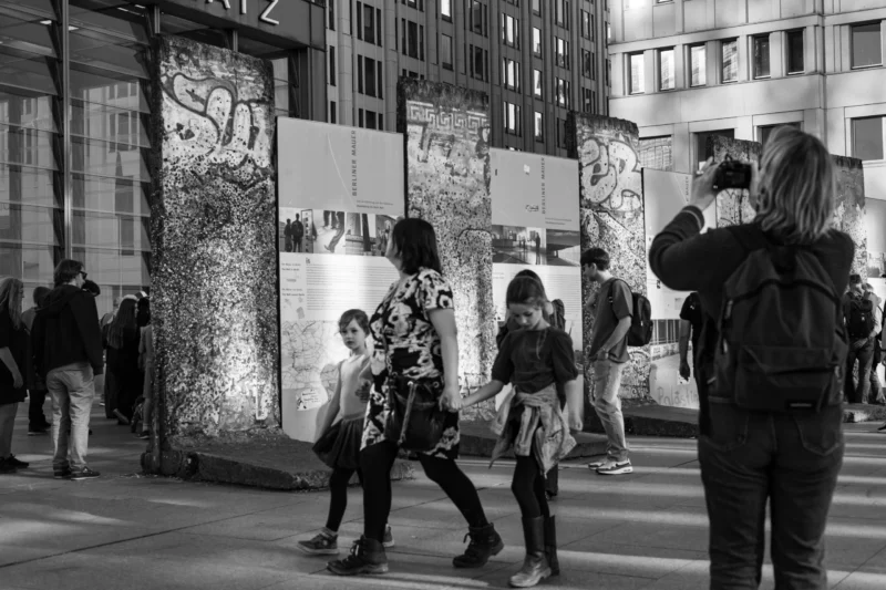 Claude · 5869 x 3913 Cette photographie en noir et blanc capture une scène urbaine animée. Le sujet principal semble être un groupe de personnes, dont une jeune fille au premier plan, vêtue d’une veste à capuche, marchant devant une fresque murale.La composition met en valeur les contrastes entre les formes architecturales et les graffitis. Le cadrage est large, incluant des passants et des bâtiments en arrière-plan. La lumière est naturelle, avec des ombres marquées, renforçant l’ambiance urbaine et dynamique. Le style est documentaire, reflétant une vie quotidienne authentique. L’ambiance est vivante et un peu mélancolique, typique des scènes de rue artistiques.