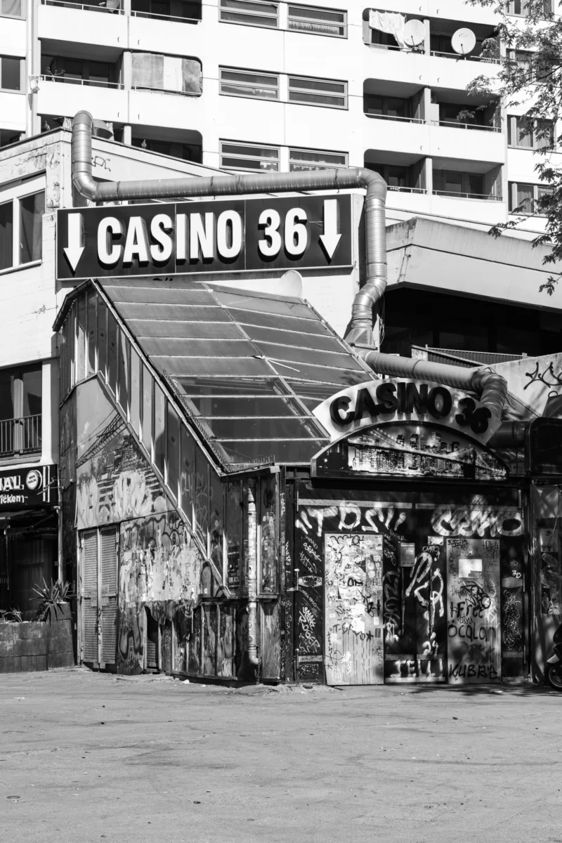 Claude · 3150 x 4725 Cette photographie en noir et blanc montre un ancien bâtiment nommé **« Casino 36 »**, partiellement dégradé. Le sujet principal est l’enseigne et la façade abîmée, évoquant un lieu abandonné.La composition met en valeur l’aspect nostalgique et décrépit, avec un cadrage serré sur l’entrée et les graffitis. La lumière, naturelle et contrastée, accentue les textures et les ombres. Les couleurs dominantes, bien que monochromes, jouent sur les nuances de gris, renforçant l’ambiance mélancolique et désuète. Le style rappelle le réalisme documentaire, créant une atmosphère à la fois mystérieuse et nostalgique.