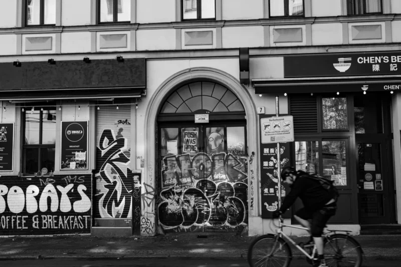 Claude · 5765 x 3843 Cette photographie en noir et blanc met en scène un cycliste passant devant une entrée d'immeuble taguée. Le sujet principal est le graffiti et l’architecture urbaine, avec des tags variés et colorés (bien que monochromes ici) sur une porte et un mur.La composition est dynamique, avec un cadrage serré sur l’entrée et une perspective légèrement basse. La lumière est naturelle, contrastée, accentuant les textures et ombres. L’ambiance est urbaine et artistique, reflétant une touche de rébellion et de vie quotidienne dans un quartier probablement animé. Le style est documentaire et esthétique, capturant l’énergie de la rue.