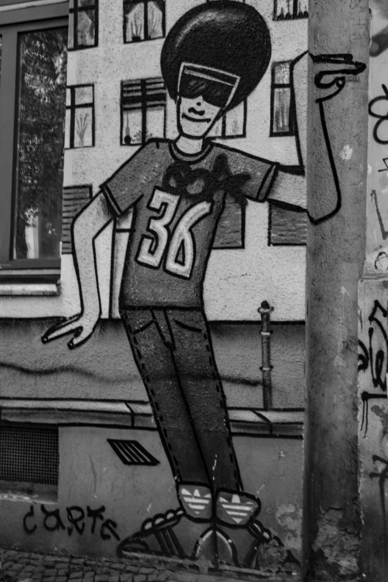 Claude · 4000 x 6000 Cette photographie montre un graffiti en noir et blanc représentant un personnage stylisé, probablement un skateur, portant un sweat à capuche avec le numéro 36 et un bonnet. Le sujet est dynamique, en posture de saut, avec des baskets et des chaussettes hautes.La composition est simple, centrée sur le personnage. Le cadrage est serré, mettant en valeur le graffiti sur un mur urbain. Les couleurs dominantes sont les nuances de gris, typiques d’une photo en noir et blanc.Le style est street art, avec des traits épais et expressifs. L’ambiance est énergique et urbaine, reflétant la culture skate et hip-hop.