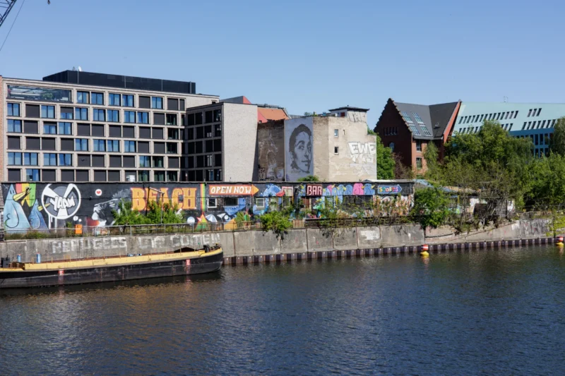 Claude · 5897 x 3931 Cette photographie montre un quartier urbain riverain. Le **sujet principal** est un ensemble de bâtiments modernes et colorés bordant un canal. La **composition** met en valeur des **graffitis** et des structures architecturales variées, avec un cadrage large incluant une partie de l’eau.La **lumière** est naturelle et claire, suggérant un jour ensoleillé. Les **couleurs dominantes** sont des tons vifs (bleus, verts, jaunes) contrastant avec le gris des bâtiments et le bleu de l’eau. Le **style** est réaliste et documentaire, capturant une ambiance **urbaine et dynamique**, mêlant art de rue et modernité architecturale.