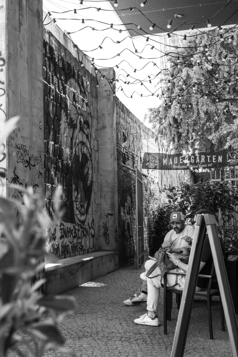 Claude · 3884 x 5826 Cette photographie en noir et blanc montre un homme assis seul sur un banc dans un espace urbain couvert, probablement une gare ou un passage souterrain. Le sujet principal est cet homme, absorbé dans ses pensées.La composition est axée sur une perspective longitudinale, avec des graffitis omniprésents sur les murs, créant une ambiance sombre et mélancolique. Le cadrage est serré, mettant en valeur l’isolement du personnage. La lumière est tamisée, renforçant l’atmosphère contemplative et un peu désenchantée. Les couleurs, bien que monochromes, accentuent les contrastes entre ombres et lumières, typiques d’un style documentaire ou street photography.
