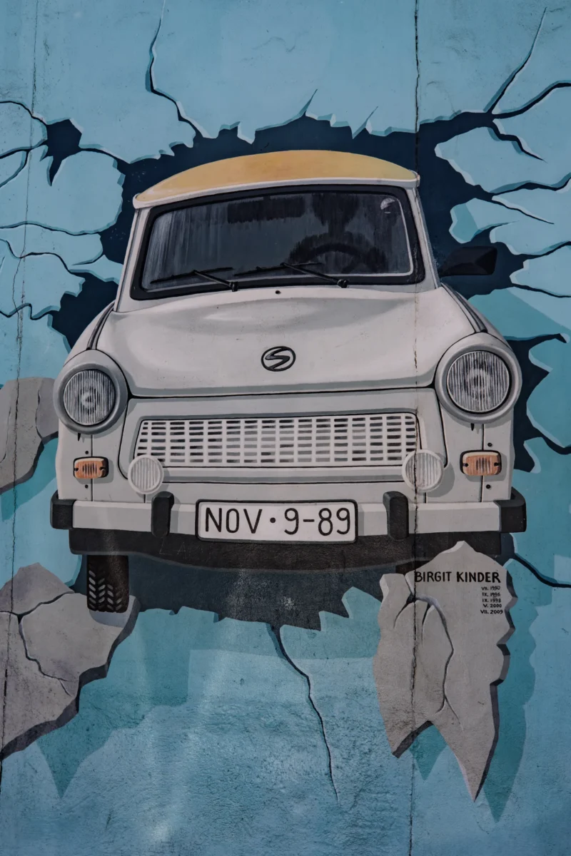 Claude · 3592 x 5388 Cette photographie met en scène une vieille voiture Trabant, modèle emblématique est-allemand, peinte en blanc avec une bande jaune sur le capot. Le sujet principal est centré, encadré par une fissure stylisée évoquant un mur brisé, donnant un effet de rupture ou de liberté.La composition est symétrique, avec un cadrage serré sur le pare-brise et la calandre. Les couleurs dominantes sont le blanc, le bleu (fond dégradé) et le jaune. La lumière semble uniforme, sans ombre marquée, créant une ambiance à la fois nostalgique et dynamique. Le style mêle réalisme et touche artistique, évoquant une esthétique à la fois rétro et moderne.
