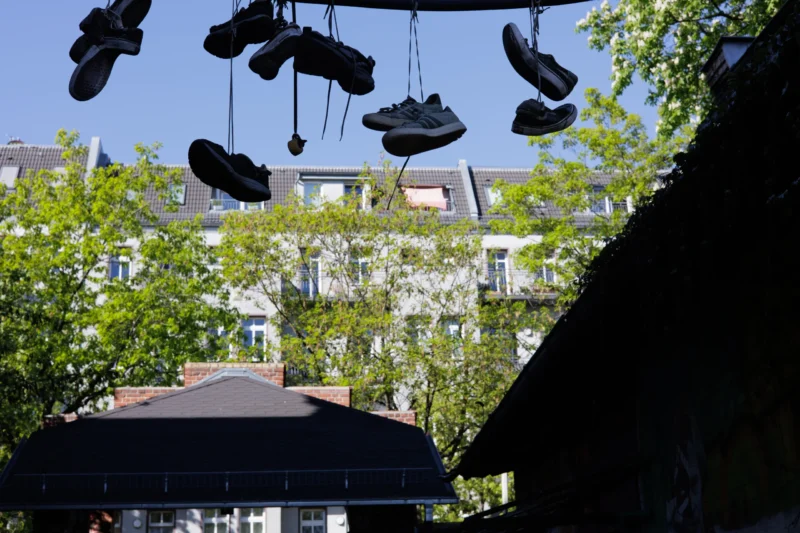 Claude · 5657 x 3771 Cette photographie met en scène une installation artistique composée de **paires de chaussures suspendues** en fil, formant un motif aérien. La **composition** joue sur le contraste entre ces éléments modernes et les bâtiments classiques en arrière-plan.Le **cadrage** est serré sur les chaussures, avec une perspective basse, tandis que l’arrière-plan montre des immeubles et des arbres. La **lumière** est naturelle et éclatante, créant des ombres nettes. Les **couleurs dominantes** sont les tons gris des chaussures, le vert des feuilles et le bleu du ciel.Le style est **urbain et poétique**, évoquant une ambiance à la fois **ludique et contemplative**.