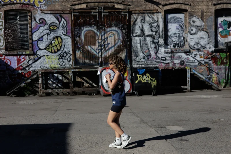 Claude · 5698 x 3799 Cette photographie met en scène une jeune femme en mouvement, marchant devant un mur tagué. Le sujet principal est dynamique, vêtue d’un short et d’un haut sportifs, avec un sac à dos.La composition joue sur la perspective avec un cadrage serré sur la personne, encadrée par le mur coloré en arrière-plan. La lumière est naturelle et vive, créant des ombres marquées. Les couleurs dominantes sont vives et contrastées : bleu, rouge, noir et blanc.Le style est urbain et spontané, évoquant une ambiance énergique et artistique, typique des rues animées et créatives.