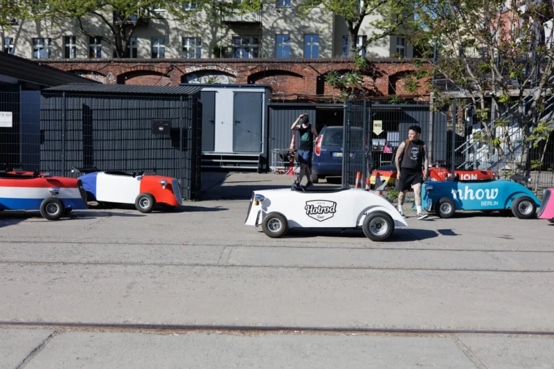 Claude · 6000 x 4000 Cette photographie montre un véhicule utilitaire blanc, probablement un golf mobile, stationné près d’un bâtiment industriel. Le sujet principal est ce véhicule, entouré de deux personnes en tenue de travail.La composition est dynamique, avec un cadrage légèrement bas, mettant en valeur le véhicule. Les rails au premier plan suggèrent un environnement urbain ou industriel. La lumière est naturelle et claire, indiquant une journée ensoleillée. Les couleurs dominantes sont les tons neutres (gris, blanc) avec des touches de bleu et de rouge sur les véhicules en arrière-plan.L’ambiance est professionnelle et moderne, évoquant un contexte de travail ou de logistique urbaine.