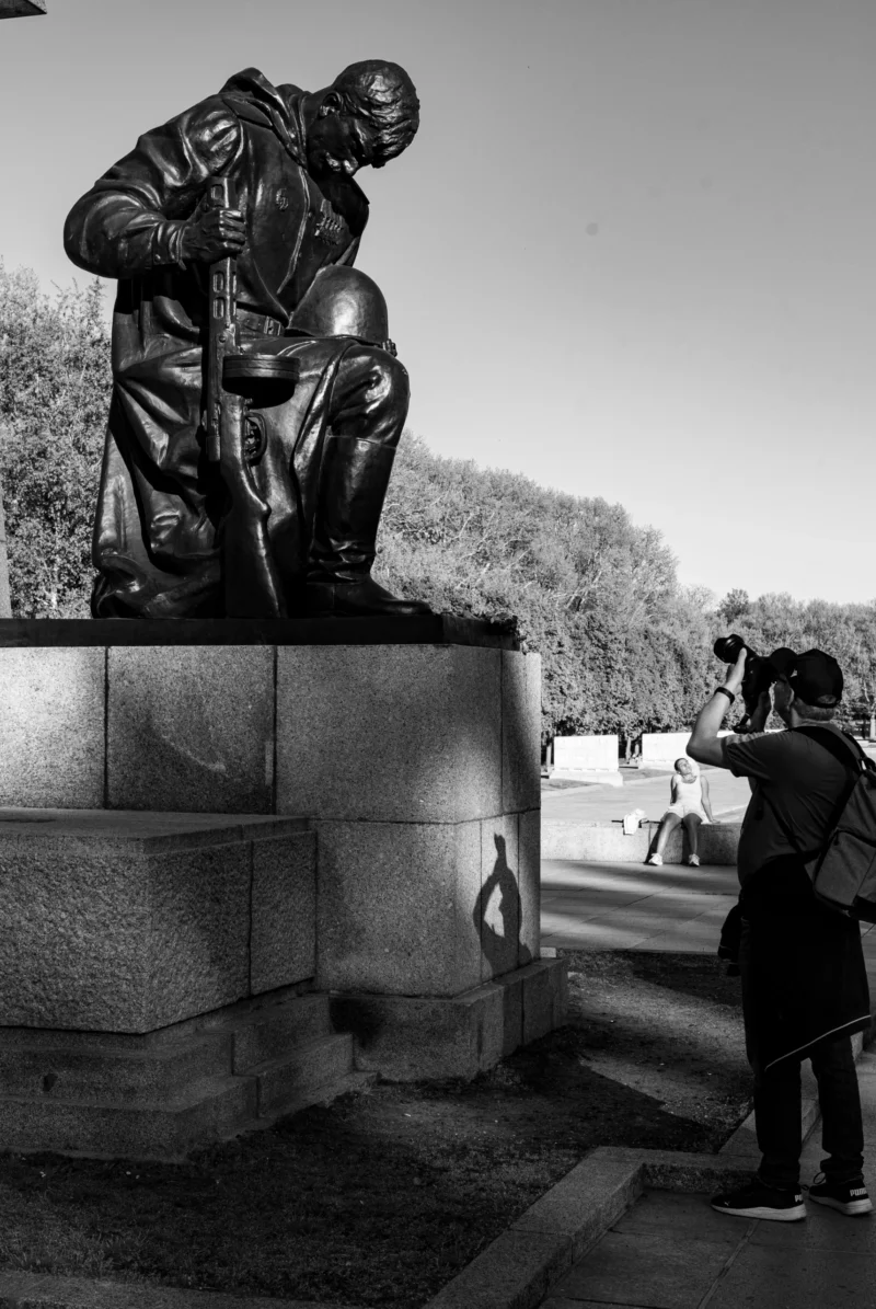 Claude · 3650 x 5451 Cette photographie en noir et blanc met en scène une statue comme sujet principal, représentant un homme assis dans une posture contemplative, les coudes sur les genoux. La composition montre la statue sur un socle, entourée d’un espace ouvert avec des arbres en arrière-plan.Le cadrage est serré sur la statue, avec une personne photographiant la scène à droite. La lumière est douce, créant des contrastes modérés. Les tons dominants sont les gris et noirs, typiques du monochrome. Le style est documentaire et intemporel, évoquant une ambiance calme et réfléchie, presque mélancolique.