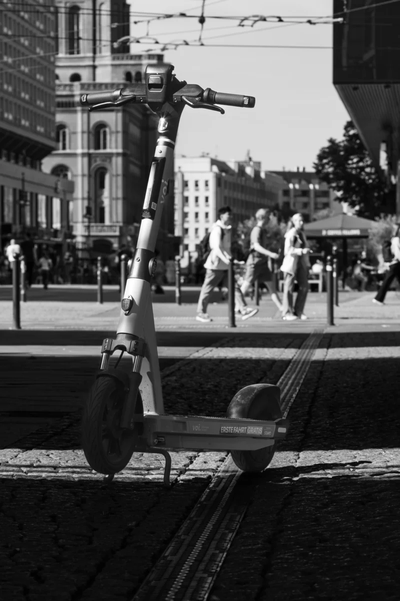 Claude · 2149 x 3224 Cette photographie en noir et blanc met en scène un scooter électrique abandonné, sujet principal, au premier plan. La composition joue sur les contrastes entre l'objet moderne et l'arrière-plan flou, où l'on distingue des passants et des bâtiments urbains.Le cadrage serré isole le scooter, tandis que l'arrière-plan suggère une ville animée. La lumière, naturelle et douce, crée des ombres marquées, renforçant le côté dramatique. Les tons dominants, du noir au gris clair, accentuent la texture des éléments.Le style est documentaire et urbain, évoquant une ambiance mélancolique et contemplative, entre modernité et quotidien.