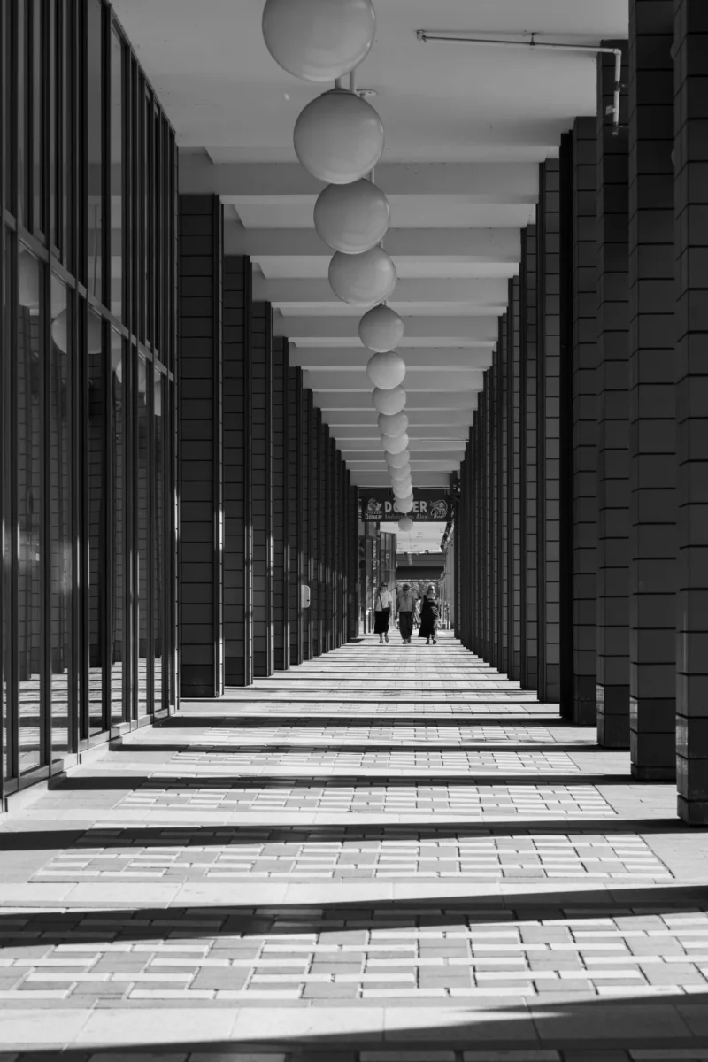 Claude · 3336 x 5004 Cette photographie en noir et blanc montre un couloir couvert bordé de vitrines et de murs en briques. Le sujet principal est l’allée centrale, symétrique et perspective, menant vers l’horizon.Le cadrage est serré, créant une profondeur avec des lignes parallèles. La lumière, naturelle et douce, accentue les contrastes entre ombres et clartés. Les couleurs dominantes, en niveaux de gris, renforcent une ambiance moderne et minimaliste.Le style épuré et architectural évoque une atmosphère à la fois froide et apaisante, presque urbaine et intemporelle.