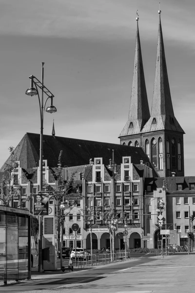 Claude · 3813 x 5720 Cette photographie met en scène une ville européenne, dominée par une cathédrale gothique à deux flèches élancées. La composition associe l’architecture religieuse imposante et des bâtiments civils aux façades ornées. Le cadrage inclut une rue bordée de lampadaires et de tables de café, créant une perspective profonde.En noir et blanc, l’image joue sur les contrastes lumineux, accentuant les textures et les ombres. Les tons clairs et foncés renforcent l’ambiance historique et solennelle, tout en évoquant une atmosphère paisible et intemporelle. Le style est documentaire et classique.