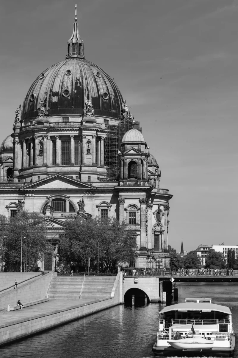 Claude · 4000 x 6000 Cette photographie met en scène la **cathédrale Saint-Edouard (Bertinskirche)** à Berlin, avec son dôme baroque comme sujet principal. La composition joue sur le contraste entre l’architecture imposante et le pont enjambant un canal, où un bateau est visible.Le cadrage est large, incluant des éléments urbains et naturels. La lumière est douce, typique d’un jour légèrement nuageux. En noir et blanc, les tons varient du clair au foncé, accentuant les textures et les volumes. Le style est documentaire et esthétique, évoquant une ambiance à la fois historique et paisible, avec une touche mélancolique.