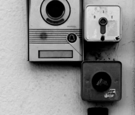 Franck T. · 09/05/2026 · 777 x 1400 Cette photographie en noir et blanc met en scène deux objets principaux : un interphone mural et un vieux téléphone portable fixé à côté. La composition est épurée, centrée sur ces éléments montés sur un mur lisse.Le cadrage serré isole ces objets, créant une ambiance minimaliste et légèrement nostalgique. La lumière est douce et uniforme, sans ombres marquées, renforçant le côté rétro. Les tons dominants sont les gris et noirs, typiques du noir et blanc, accentuant le style vintage et intemporel. L’ambiance évoque la simplicité et la technologie ancienne.