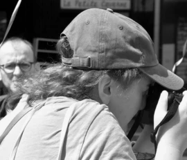 Franck T. · 09/05/2026 · 1400 x 771 Cette photographie en noir et blanc met en scène une personne comme sujet principal, portant un chapeau et tenant un appareil photo devant son œil. La composition montre aussi un homme à l’arrière-plan, légèrement flou.Le cadrage est serré sur le visage principal, avec une profondeur de champ réduite. La lumière semble naturelle, douce et diffuse, créant des contrastes modérés. L’ambiance est intime et spontanée, évoquant un moment de capture photographique.Le style est documentaire et réaliste, renforcé par l’absence de couleurs.