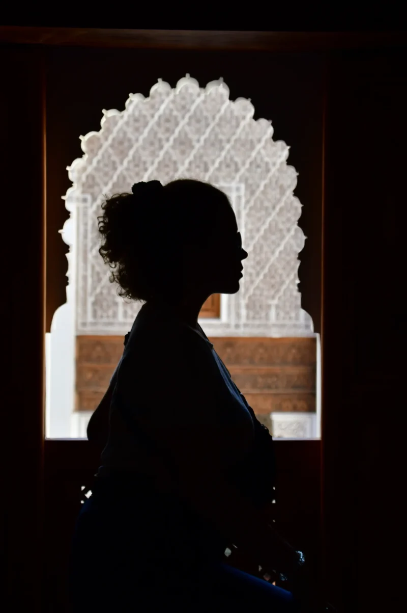 Vincent · 14/10/2022 · 3215 x 4834 Cette photographie met en scène une silhouette féminine en contre-jour, sujet principal. La composition repose sur un cadrage serré, avec un jeu d’ombres et de lumière accentué par une fenêtre décorée de motifs ajourés.La lumière, douce et tamisée, crée un effet dramatique, tandis que les couleurs dominantes sont les tons sombres (noirs, bleus profonds) contrastant avec les blancs des motifs lumineux. Le style évoque une ambiance poétique et introspective, presque onirique, renforcée par la texture des éléments architecturaux. L’atmosphère est à la fois mystérieuse et élégante.