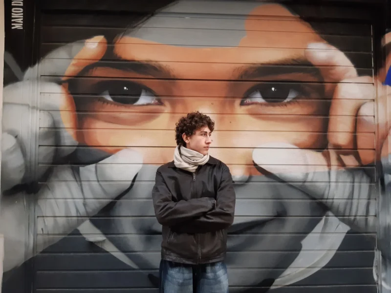 Vincent · 28/01/2024 · 4608 x 3456 Cette photographie met en scène un jeune homme comme sujet principal, debout devant un mural. La composition joue sur le contraste entre son visage et celui du personnage peint derrière lui, tous deux aux yeux expressifs. Le cadrage est serré, centré sur le sujet, avec une légère profondeur de champ.La lumière est douce, naturelle, mettant en valeur les traits du jeune homme et les détails du mural. Les couleurs dominantes sont les tons froids (gris, noir) et chauds (orange, brun) du mural, créant une ambiance à la fois urbaine et artistique. Le style est réaliste et visuellement percutant, évoquant une atmosphère à la fois contemplative et dynamique.