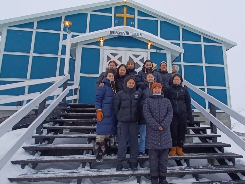 Pierre · 25/02/2024 · 1872 x 1404 Cette photographie montre un groupe de personnes posant devant une église enneigée. Le sujet principal est ce groupe, composé majoritairement de femmes, vêtues chaudement.La composition met en avant les marches menant à l’entrée de l’église, encadrée par des vitraux bleus. Le cadrage est serré sur le groupe, avec une lumière naturelle diffuse, typique d’un jour hivernal.Les couleurs dominantes sont les bleus de l’église, le blanc de la neige et les tons sombres des vêtements. Le style est documentaire et chaleureux, créant une ambiance conviviale et paisible malgré le froid.