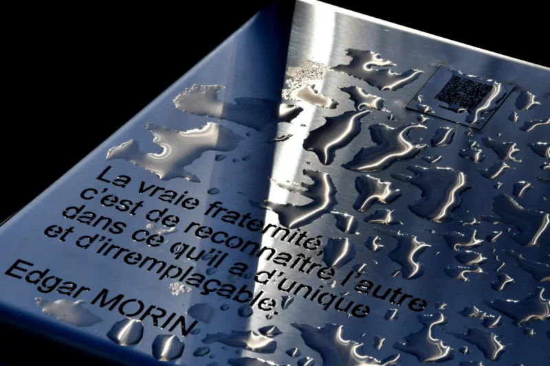 Vincent · 17/01/2026 · 5400 x 3600 Cette photographie met en scène une plaque gravée avec une citation d’Edgar Morin. Le sujet principal est le texte en français, partiellement caché par une surface glacée ou métallique réfléchissante.La composition joue sur le contraste entre le texte lisible et la texture fracturée et brillante de la surface. Le cadrage est serré, centré sur la plaque.La lumière est douce mais met en valeur les reflets et les détails. Les couleurs dominantes sont les tons froids (bleutés, gris) et le blanc du texte.Le style est épuré et artistique, créant une ambiance à la fois intellectuelle et mystérieuse.