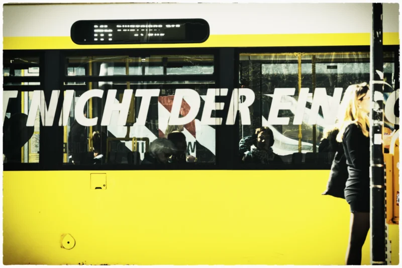 Thierry · 30/04/2026 · 3840 x 2560 Cette photographie montre un tram jaune avec un graffiti partiel en lettres rouges et blanches ("WIR SIND DIE..."). Le sujet principal est le graffiti et les passagers à l'intérieur du tram.La composition est dynamique, avec un cadrage serré sur le côté du tram. La lumière est tamisée, créant des contrastes doux. Les couleurs dominantes sont le jaune vif du tram et les tons rouges/blancs du graffiti. L’ambiance est à la fois urbaine et légèrement mystérieuse, renforcée par les silhouettes floues des passagers. Le style est documentaire et réaliste.