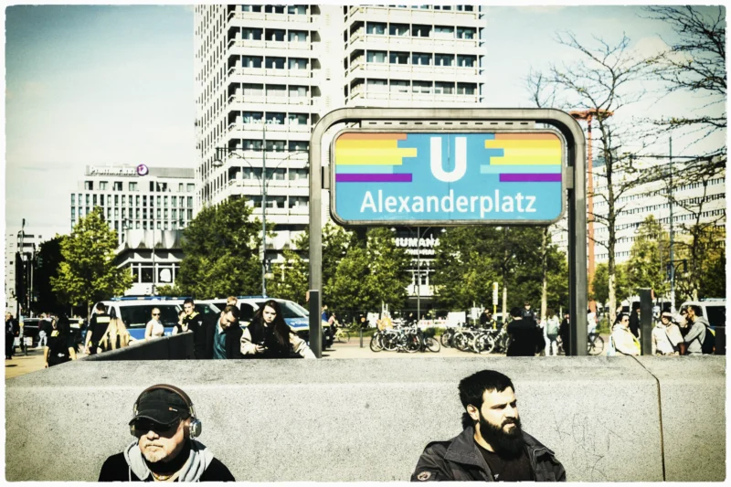 Thierry · 30/04/2026 · 3840 x 2560 Cette photographie montre un panneau indiquant "U Alexanderplatz" avec des couleurs vives (bleu, jaune, rouge). Le sujet principal est le panneau et les deux hommes en premier plan, assis et regardant vers la gauche.La composition est dynamique, avec une foule en arrière-plan. Le cadrage est serré sur les personnages et le panneau, intégrant aussi des bâtiments et des arbres. La lumière semble naturelle, avec des contrastes doux. Les couleurs dominantes sont les teintes vives du panneau et les tons neutres de la ville.Le style est réaliste et urbain, évoquant une ambiance quotidienne et animée.