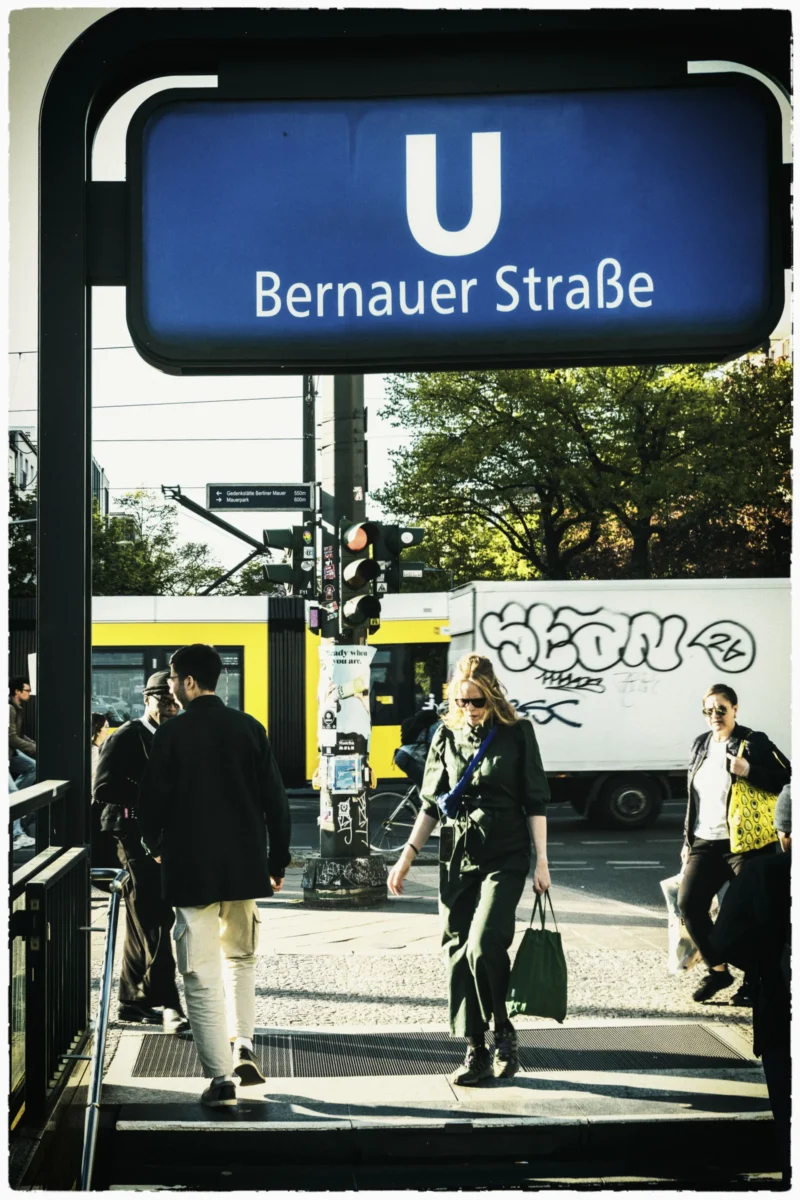 Thierry · 30/04/2026 · 2560 x 3840 Cette photographie montre un arrêt de tramway à Bernauer Straße. Le sujet principal est un panneau indicateur "U" (métro) avec le nom de la station. La composition inclut des passagers en mouvement, un tramway jaune et un bus blanc en arrière-plan. Le cadrage est serré sur le panneau et les personnes, avec une profondeur de champ moyenne.La lumière est naturelle et semble provenir d’un jour ensoleillé. Les couleurs dominantes sont les tons gris, bleu du panneau, jaune du tramway et blanc du bus. Le style est documentaire et réaliste, capturant une scène urbaine quotidienne. L’ambiance est dynamique et typique d’une ville active.