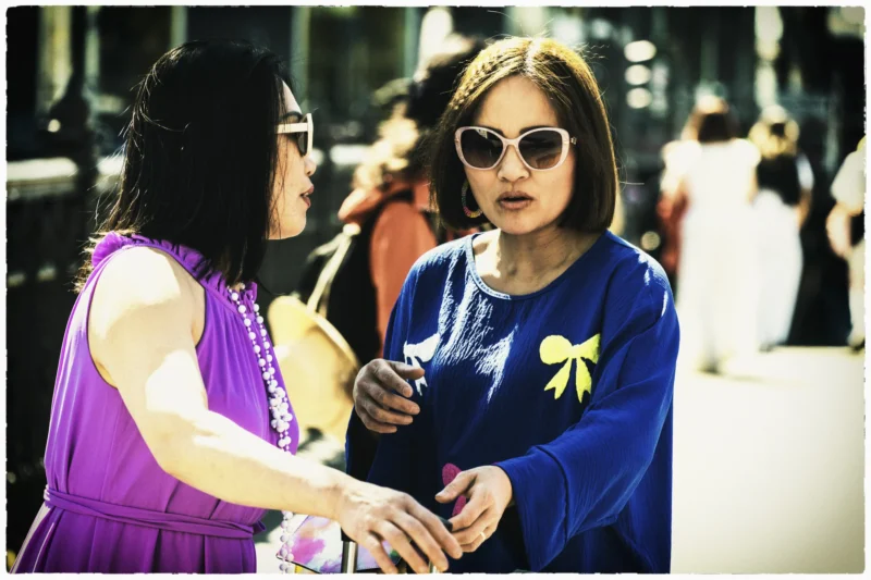 Thierry · 01/05/2026 · 3840 x 2560 Cette photographie met en scène deux femmes comme sujets principaux. L’une, vêtue d’une tenue pourpre et accessoirisée de fleurs hawaïennes, semble engagée dans une conversation avec l’autre, habillée d’un pull bleu et portant des lunettes de soleil.Le cadrage est serré, centré sur leurs visages et leurs mains, créant une ambiance intime. La lumière semble naturelle, avec des contrastes doux. Les couleurs dominantes sont vives : violet, bleu et touches de blanc.Le style évoque un mélange de spontanéité et de nostalgie, renforcé par le flou des éléments arrière-plan. L’ambiance est chaleureuse et légèrement théâtrale.