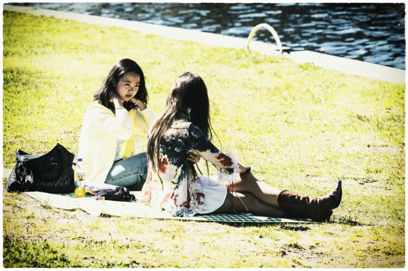 Thierry · 01/05/2026 · 3840 x 2560 Cette photographie montre deux jeunes femmes assises sur une couverture dans un parc, près d’un plan d’eau. Le sujet principal est leur interaction détendue, avec une ambiance conviviale.La composition est naturelle, avec un cadrage serré sur les deux personnes. La lumière, douce et légèrement dorée, suggère un moment en fin de journée. Les couleurs dominantes sont des tons naturels : verts pour l’herbe, beiges et blancs pour les vêtements, avec des touches de bleu pour l’eau.Le style est simple et spontané, évoquant un instant de vie quotidienne. L’ambiance est calme et chaleureuse, presque nostalgique grâce au filtre sépia appliqué.