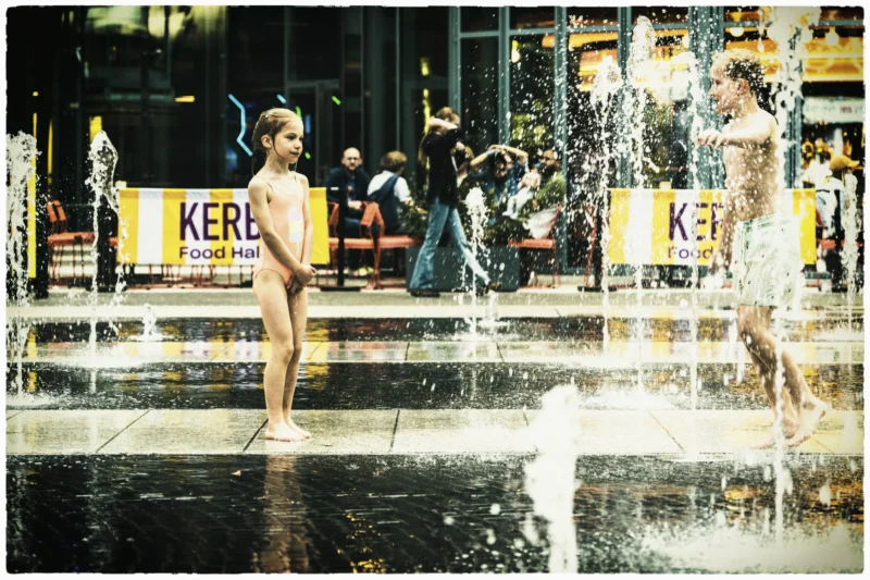 Thierry · 01/05/2026 · 3840 x 2560 Cette photographie capture deux enfants jouant dans des jets d’eau. Le sujet principal est leur insouciance et leur joie, contrastant avec l’environnement urbain.La composition est dynamique, avec un cadrage serré sur les enfants au premier plan, tandis que l’arrière-plan montre des passants et des enseignes "Kerplop". La lumière semble naturelle, avec des ombres douces, créant une ambiance lumineuse et estivale. Les couleurs dominantes sont des tons chauds et clairs, avec des reflets argentés de l’eau.Le style est documentaire et spontané, évoquant une ambiance joyeuse et détendue.
