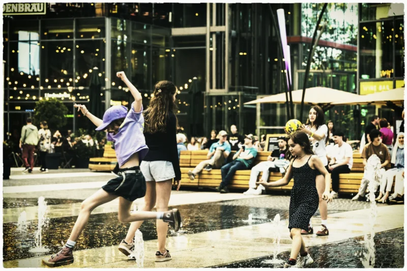 Thierry · 01/05/2026 · 3840 x 2560 Cette photographie capture un moment festif en extérieur autour d’une fontaine. Le sujet principal est un groupe de jeunes gens, dont une femme levant les bras, semblant danser ou célébrer.La composition est dynamique, avec des personnes en mouvement et un cadrage serré sur elles. L’arrière-plan montre des spectateurs assis et des structures urbaines.La lumière est douce et légèrement dorée, évoquant un coucher de soleil. Les couleurs dominantes sont des tons chauds (oranges, bruns) et des teintes pastel, avec des éclairages artificiels ajoutant une ambiance festive.Le style est spontané et naturel, créant une ambiance joyeuse et conviviale.