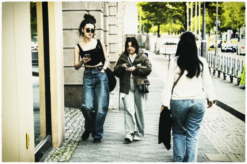 Thierry · 01/05/2026 · 3840 x 2560 Cette photographie capture trois jeunes femmes en mouvement sur un trottoir urbain. Le sujet principal semble être la femme au centre, vêtue d’un jean et d’un haut sombre, tenant un objet à la main.La composition est dynamique, avec un cadrage serré sur les personnages et un arrière-plan flou. La lumière, naturelle et douce, donne une ambiance tamisée, typique d’un jour légèrement nuageux. Les couleurs dominantes sont des tons neutres (gris, beige) avec des touches de bleu et de vert, renforçant une atmosphère moderne et décontractée.Le style est réaliste et spontané, évoquant une scène de vie quotidienne. L’ambiance est à la fois urbaine et intimiste.