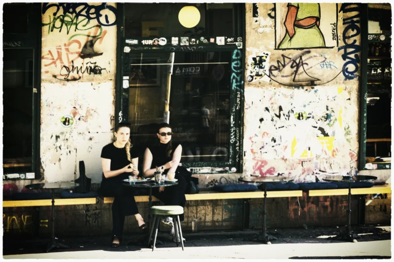 Thierry · 02/05/2026 · 3840 x 2560 Cette photographie montre deux personnes assises derrière un comptoir dans un espace graffití et coloré, probablement un café ou un lieu artistique. Le sujet principal semble être l’ambiance créative et décontractée.La composition est symétrique avec les deux individus au centre. Le cadrage est serré, mettant en valeur les détails des murs couverts de tags et des objets autour. La lumière est tamisée, créant une ambiance chaleureuse et intimiste. Les couleurs dominantes sont vives et contrastées, avec des tons de jaune, orange, noir et blanc.Le style rappelle l’art urbain, dynamique et spontané, évoquant une ambiance bohème et artistique.