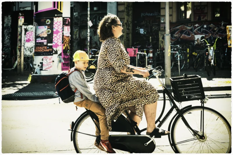 Thierry · 02/05/2026 · 3840 x 2560 Cette photographie met en scène deux femmes à vélo en ville. Le sujet principal est la femme assise à l’arrière, vêtue d’une robe à motifs léopard, tandis que la passagère, plus jeune, porte un chapeau jaune.La composition est dynamique, avec un cadrage serré sur les deux personnages. La lumière est naturelle, douce et légèrement contrastée, créant des ombres nettes. Les couleurs dominantes sont les tons chauds (jaune, marron) et les teintes pastel.Le style est documentaire et spontané, évoquant une ambiance urbaine détendue et estivale.