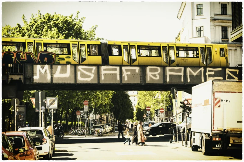 Thierry · 02/05/2026 · 3840 x 2560 Cette photographie montre un tram suspendu au-dessus d’une rue avec un graffiti "MUSEUM BAR" sur un panneau. Le sujet principal est ce tram jaune et le graffiti.La composition est dynamique, avec une perspective en contre-plongée. Le cadrage inclut des éléments urbains comme des voitures, des passants et des bâtiments.La lumière est douce, typique d’un jour ensoleillé, créant des ombres légères. Les couleurs dominantes sont les tons jaunes, gris et verts.Le style est documentaire, avec une touche vintage due au filtre sépia. L’ambiance est urbaine et vivante, mêlant mouvement et vie quotidienne.