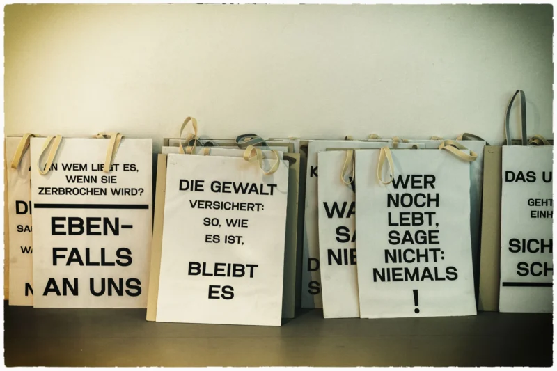Thierry · 02/05/2026 · 3840 x 2560 Cette photographie montre plusieurs sacs en papier suspendus, portant des slogans politiques en allemand. Le sujet principal est ces messages engagés, évoquant des thèmes comme la violence, la responsabilité et la non-violence.La composition est horizontale et serrée, avec un cadrage centré sur les sacs. La lumière est douce et uniforme, sans ombres marquées. Les couleurs dominantes sont les tons neutres (beige, blanc) des sacs, rehaussés par les inscriptions en noir.Le style est épuré et direct, créant une ambiance sobre et militante, soulignant l'urgence des messages portés.