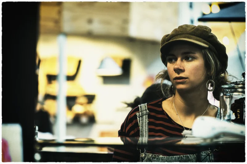 Thierry · 02/05/2026 · 3840 x 2560 Cette photographie met en scène une jeune femme comme sujet principal, assise dans un espace industriel ou artisanal. Elle porte un bonnet et une veste à carreaux, tout en tenant un objet près de son visage.La composition est centrée sur elle, avec un cadrage serré qui isole son visage et ses mains. L’arrière-plan flou révèle des éléments techniques et des couleurs chaudes.Les couleurs dominantes sont les tons bruns et gris, adoucis par des éclairages doux. La lumière semble naturelle, créant une ambiance à la fois réaliste et légèrement mélancolique, évoquant un style documentaire ou cinématographique intimiste.
