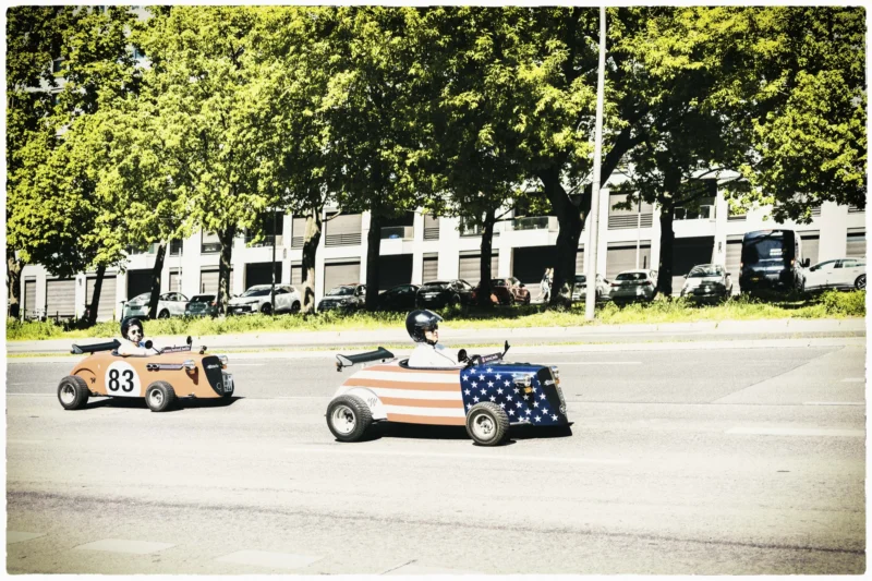 Thierry · 02/05/2026 · 3840 x 2560 Cette photographie capture deux petites voitures de course vintage en compétition sur une route. Le sujet principal est ces voitures, numérotées 83 et 84, avec des designs rétro.La composition montre un cadrage dynamique, centré sur les véhicules en mouvement. L’arrière-plan, avec des bâtiments et des arbres, est flou, accentuant le sujet. La lumière semble naturelle, avec des tons doux et une ambiance légèrement nostalgique.Les couleurs dominantes sont des teintes neutres et terreuses, avec des touches de bleu et de rouge sur la voiture 84. Le style évoque une ambiance vintage et sportive, renforcée par le flou artistique et les tons sépia.