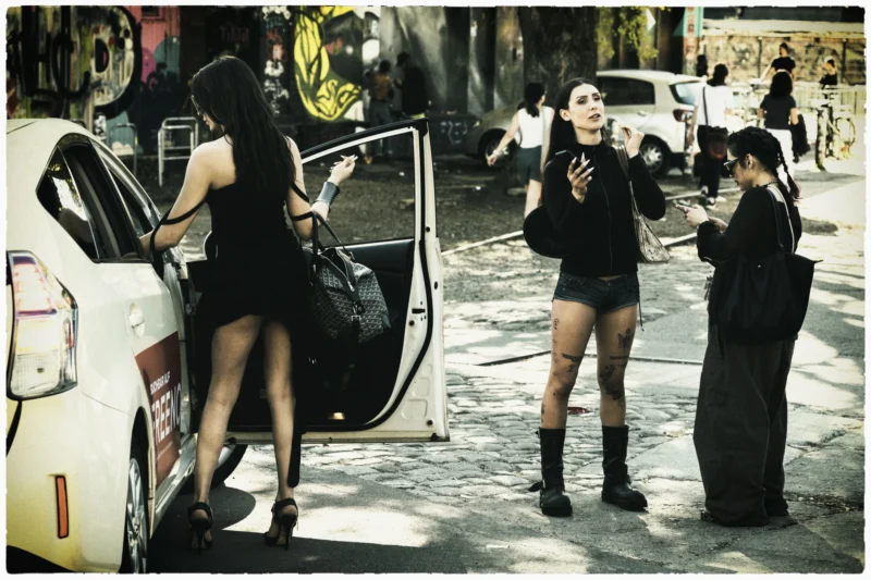 Thierry · 02/05/2026 · 3840 x 2560 Cette photographie capture trois jeunes femmes en milieu urbain. Le sujet principal semble être la femme au centre, vêtue d’une tenue courte et noire, en train de parler au téléphone. La composition montre une scène dynamique avec des éléments de rue en arrière-plan.Le cadrage est serré sur les personnages, avec une profondeur de champ réduite. La lumière est naturelle, légèrement contrastée, créant des ombres marquées. Les couleurs dominantes sont des tons sombres (noir, gris) avec des touches de blanc (voiture) et des détails colorés (graffiti, vêtements).Le style évoque un réalisme brut, typique d’une photographie documentaire ou street style. L’ambiance est à la fois moderne et spontanée, reflétant une vie urbaine animée.