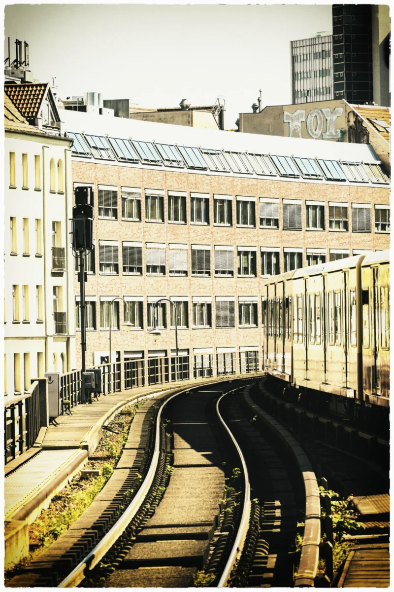 Thierry · 03/05/2026 · 2560 x 3840 Cette photographie met en scène une voie ferrée urbaine en forme de boucle, sujet principal. La composition montre des bâtiments anciens en arrière-plan, encadrés par des rails métalliques et des poteaux.Le cadrage est serré, centré sur les rails et les structures environnantes. La lumière est douce et naturelle, avec une teinte légèrement sépia, évoquant une ambiance nostalgique. Les couleurs dominantes sont les tons gris-beige, adoucis par des touches de vert et de bleu.Le style est documentaire et légèrement vintage, créant une ambiance mélancolique et industrielle.