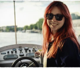 Thierry · 07/05/2026 · 3840 x 2560 Cette photographie met en scène une jeune femme aux cheveux roux, sujet principal, souriante et portant des lunettes de soleil. Elle est assise au volant d’un bateau, avec une composition centrée sur son visage et ses mains sur le volant.Le cadrage est serré, mettant en valeur son expression joyeuse. La lumière est douce et naturelle, probablement en extérieur par temps clair. Les couleurs dominantes sont les tons chauds (roux, bleu marine de sa veste) et les teintes pastel du bateau et de l’eau.Le style est décontracté et spontané, évoquant une ambiance détendue et estivale.