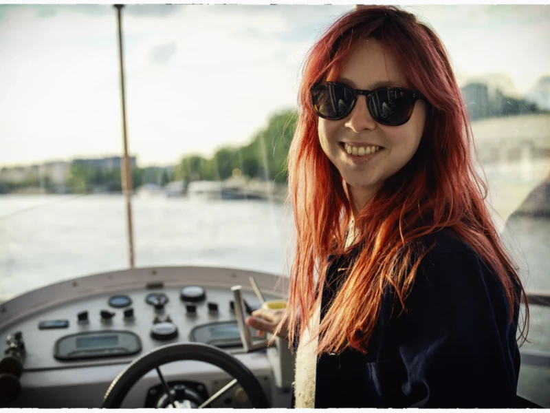 Thierry · 07/05/2026 · 3840 x 2560 Cette photographie met en scène une jeune femme aux cheveux roux, sujet principal, souriante et portant des lunettes de soleil. Elle est assise au volant d’un bateau, avec une composition centrée sur son visage et ses mains sur le volant.Le cadrage est serré, mettant en valeur son expression joyeuse. La lumière est douce et naturelle, probablement en extérieur par temps clair. Les couleurs dominantes sont les tons chauds (roux, bleu marine de sa veste) et les teintes pastel du bateau et de l’eau.Le style est décontracté et spontané, évoquant une ambiance détendue et estivale.