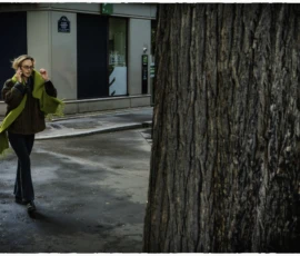 Thierry · 16/05/2026 · 3840 x 2560 Cette photographie met en scène une femme comme sujet principal, vêtue d’un manteau vert clair, prenant une photo avec son téléphone. La composition joue sur le contraste entre la silhouette humaine et le tronc d’arbre texturé à droite. Le cadrage serré isole le sujet dans un espace urbain, avec une porte en arrière-plan.La lumière, tamisée et légèrement bleutée, suggère un crépuscule ou un éclairage artificiel. Les couleurs dominantes sont les tons froids (gris, bleu) et le vert du manteau, créant une ambiance calme et introspective. Le style est sobre et réaliste, évoquant une atmosphère contemplative et moderne.