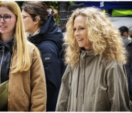 Thierry · 16/05/2026 · 3840 x 2560 Cette photographie met en scène une femme au premier plan comme sujet principal, avec des cheveux bouclés et un manteau clair. Elle semble marcher en extérieur.La composition montre aussi deux autres personnes en arrière-plan, légèrement floues. Le cadrage est serré sur les visages et le haut du corps.La lumière est naturelle et semble tamisée, peut-être par un temps nuageux. Les couleurs dominantes sont les tons neutres (beige, gris) avec des touches de vert et de bleu en arrière-plan.Le style est documentaire et spontané, capturant une scène de vie quotidienne. L’ambiance est sobre et réaliste.