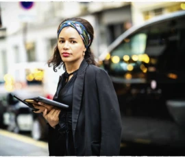 Thierry · 15/05/2026 · 3840 x 2560 Cette photographie met en scène une femme comme sujet principal, vêtue d’un manteau noir et tenant une tablette. La composition est simple et centrée, avec un cadrage serré sur elle, légèrement décentré vers la gauche.La lumière est douce et naturelle, probablement en fin de journée, créant des contrastes subtils. Les couleurs dominantes sont les tons sombres (noir, gris) avec des éclats chauds (lueurs des enseignes et phares de voitures).Le style est sobre et urbain, évoquant une ambiance moderne et professionnelle, tout en capturant une touche de mélancolie.