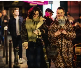Thierry · 15/05/2026 · 3840 x 2560 Cette photographie capture deux femmes marchant de front sur un trottoir urbain la nuit. Elles constituent le sujet principal, habillées de manière stylisée : l’une porte un manteau vert foncé et l’autre un long manteau à motifs léopard.La composition est dynamique, avec un cadrage serré sur elles, entourées d’une foule floue en arrière-plan. La lumière, tamisée et artificielle, crée une ambiance nocturne et chaleureuse. Les couleurs dominantes sont les tons sombres (noir, vert, marron) adoucis par des éclairages chauds.Le style est urbain et contemporain, évoquant une ambiance moderne et animée.